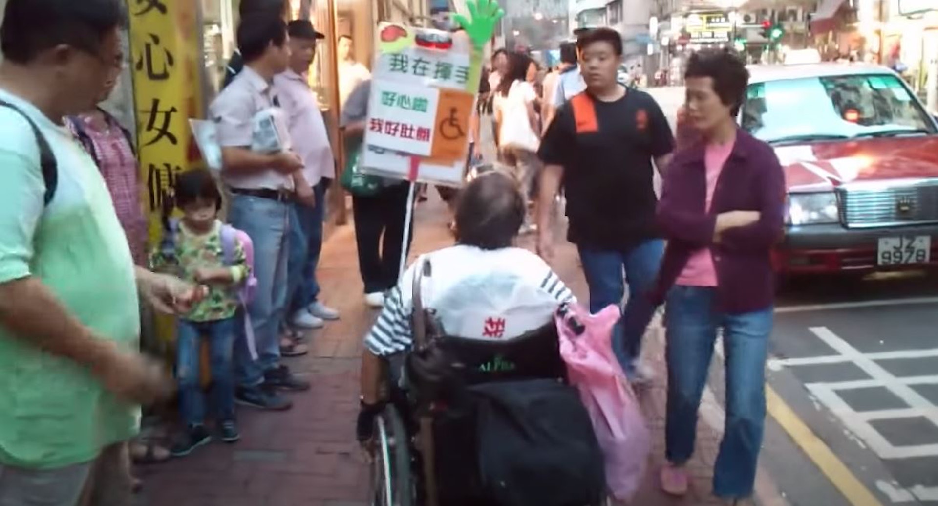 「安蔭婆婆」離開的士車廂坐上輪椅，其輪椅上插上一個寫有「我在揮手」、「請前來吧」和「我想坐車」的字句，而且印有一個輪椅人士圖案，字牌背後寫上「我好肚餓」。（網上影片截圖）